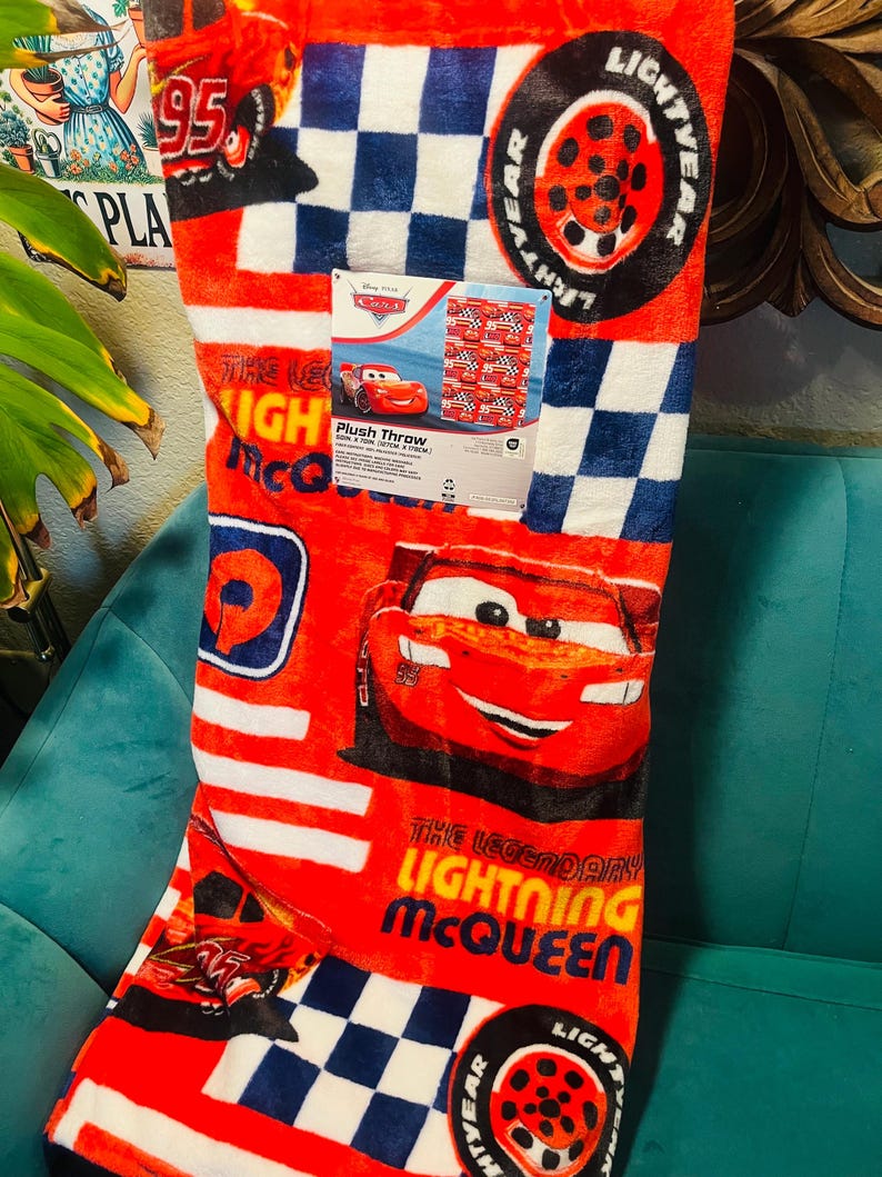 Puede incluir: Manta de felpa roja con Lightning McQueen de la pel&iacute;cula Cars. La manta tiene un patr&oacute;n de cuadros en blanco y azul marino, con gr&aacute;ficos de Lightning McQueen y neum&aacute;ticos. Tambi&eacute;n es visible el texto "The Legendary Lightning McQueen".