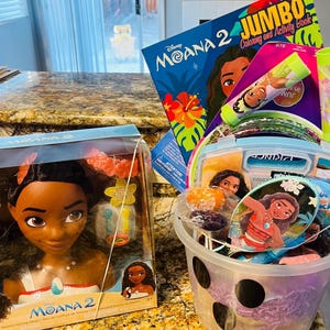 Puede incluir: Una cesta de regalo llena de art&iacute;culos tem&aacute;ticos de Moana. Incluye una mu&ntilde;eca de cabeza de peinado de Moana, un libro para colorear y actividades, una varita de burbujas y otros accesorios. La cesta es morada y blanca.