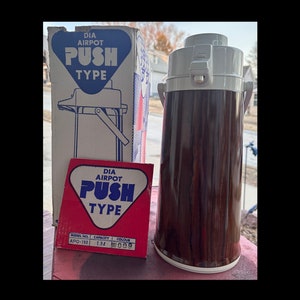 May include: A vintage DIA Airpot push-type thermos with a wood-grain finish, alongside its original box and a smaller red label. The thermos has a white top and handle. The box features the same logo and product illustration.