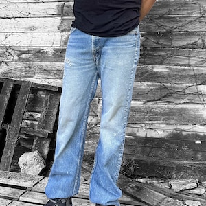May include: A pair of light blue denim jeans with a faded and distressed look. The jeans have a straight leg fit and are worn by a person standing in front of a wooden wall.