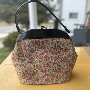 Puede incluir: Un bolso vintage de mano con estampado floral, parte superior de charol negro y cierre dorado. El bolso tiene un asa corta.