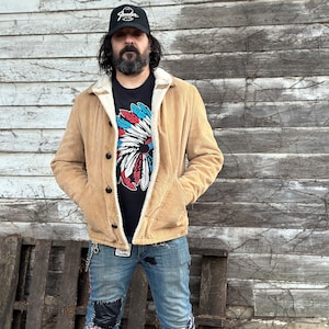 May include: A man wearing a tan corduroy jacket with a sherpa lining, a black t-shirt with a colorful Native American headdress design, blue jeans, and a black baseball cap with the word "Fender" on it.