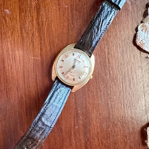 May include: A vintage gold-toned wristwatch with a black leather strap. The watch face is light-coloured with black numerals and hands. The watch is set against a wooden surface.