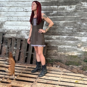 Puede incluir: Un vestido lencero marrón con ribete de encaje blanco y un corpiño decorativo. El vestido se combina con botas de combate negras. El fondo presenta una pared de madera desgastada y un palé de madera.