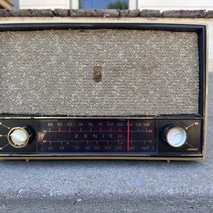 May include: A vintage Zenith radio with a black and beige color scheme. The radio has a fabric speaker grille, a dial with numbers from 55 to 160, and a knob for adjusting the volume. The radio is sitting on a gray concrete surface.