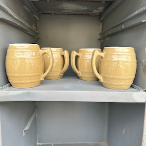 May include: Four beige ceramic mugs with handles sit on a gray shelf inside a metal cabinet.