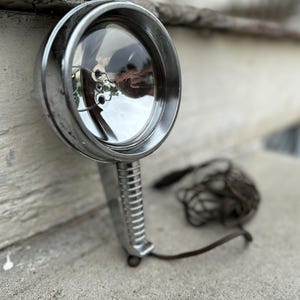 May include: A vintage chrome spotlight with a ribbed handle and a clear lens. The spotlight is resting on a concrete surface, with a wire mesh object and a cord nearby. The overall aesthetic is industrial.