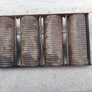 Op de afbeelding: Een antieke metalen broodvorm met vier cilindrische compartimenten. De vorm en cilinders hebben een roestig, verouderd uiterlijk, wat wijst op vintage gebruik. Het ontwerp omvat een rechthoekig frame met verticale verdelers.