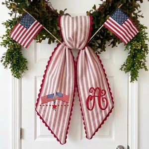 May include: A patriotic door decoration featuring a green wreath, two American flags, and a red and white striped fabric bow. The bow has a scalloped edge and embroidered details, including an initial and flags.