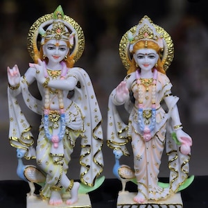 May include: Two white statues of deities, decorated with gold and intricate details. Each figure wears a crown and draped clothing, with one holding a flute. The statues stand on decorative bases with peacock figures.