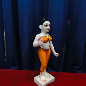 May include: A white marble statue of a person in a standing pose. The figure wears an orange top and leggings with gold accents. The statue is on a white square base, with a red surface and blue curtain backdrop.