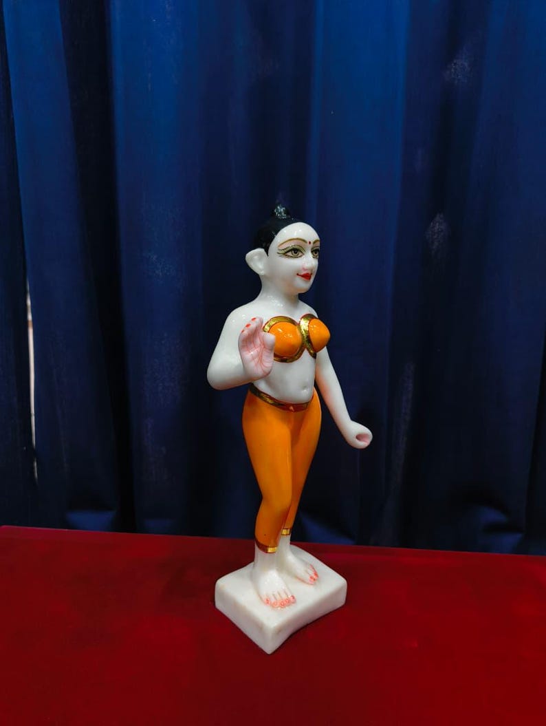 May include: A white marble statue of a person in a standing pose. The figure wears an orange top and leggings with gold accents. The statue is on a white square base, with a red surface and blue curtain backdrop.