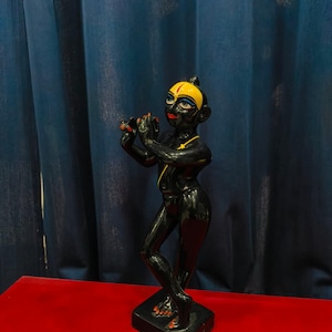 May include: A black statue of a figure playing a flute, with yellow and red accents. The figure has a yellow head covering and red lips. It stands on a black base, set against a blue curtain and a red surface.