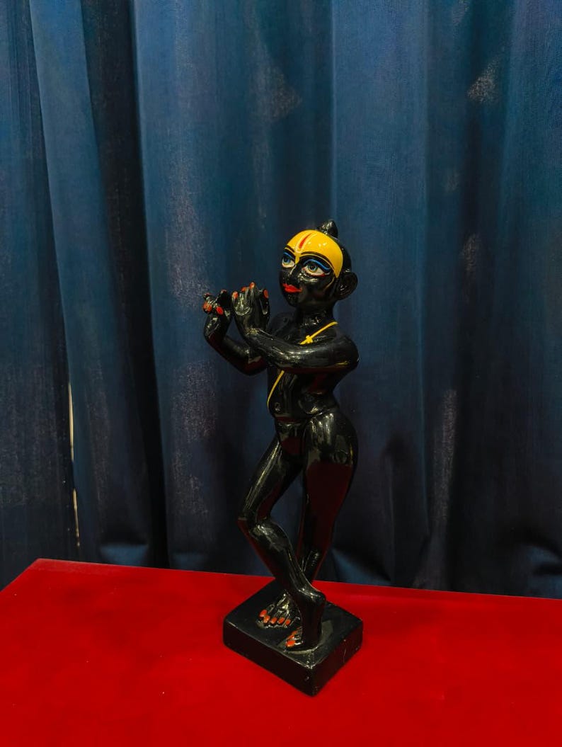 May include: A black statue of a figure playing a flute, with yellow and red accents. The figure has a yellow head covering and red lips. It stands on a black base, set against a blue curtain and a red surface.