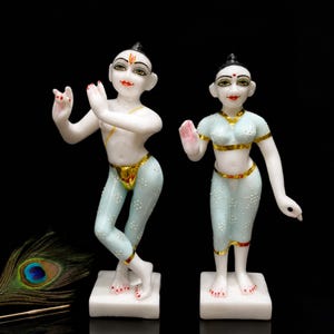 May include: Two white marble statues of figures in traditional attire. One figure is in a dancing pose, the other is standing with a hand raised. Both figures have gold accents and are on white square bases.