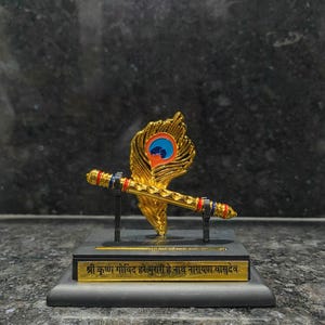 May include: A gold-colored decorative piece featuring a flute and a peacock feather design. The flute has red and blue accents. The base has gold text on a black background. The text reads "श्री कृष्ण गोविंद हरे मुरारी हे नाथ नारायण वासुदेव".
