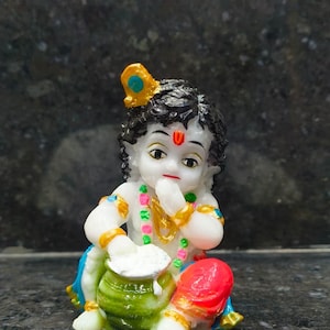 Peut inclure: Une petite figurine d'enfant aux cheveux noirs, ornée d'une plume de paon dorée et d'une marque rouge sur le front. Le personnage a la peau blanche, porte des colliers de perles colorés et des bracelets dorés, et tient un pot vert de beurre blanc.