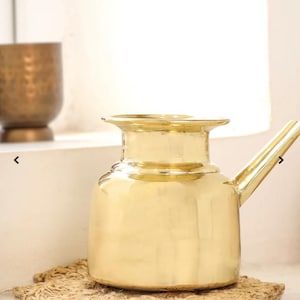 May include: A gold-colored metal pitcher with a long spout and a wide, rounded body. The pitcher sits on a woven mat, with a matching gold-colored cup in the background. The pitcher has a classic, elegant design.