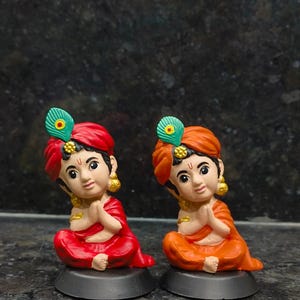 Könnte beinhalten: Zwei Figuren einer sitzenden Figur mit gefalteten Händen. Eine Figur trägt einen roten Turban und ein rotes Outfit, die andere einen orangenen Turban und ein orangefarbenes Outfit. Beide haben goldene Ohrringe und eine Pfauenfeder. Sie sitzen auf einem schwarzen Sockel.