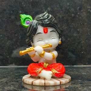 May include: A small figurine of a deity with white skin, black hair, and a red mark on the forehead. The figure wears bright red pants with gold trim, holds a gold flute to the lips, and features a green and purple peacock feather in the hair.