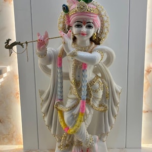 May include: A white marble statue of a deity standing in a crossed-leg pose, playing a thin gold flute. The figure wears a pink turban with a peacock feather, gold jewellery, and a multi-coloured garland. The base is rectangular with gold detailing.