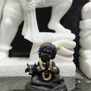 以下が含まれることがあります： 祈りのポーズで座っている黒い像。金色のネックレスと金の頭飾りを身につけています。小さな黒い牛が神の左側にいます。像は濃い灰色の台座にあり、背景には白い像があります。