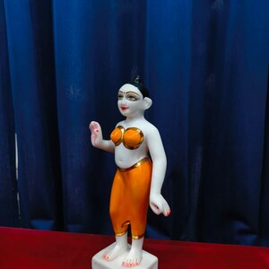 May include: A white and orange figurine of a person with black hair and red lips. The figurine is wearing an orange top and pants with gold trim. The figurine is standing on a white square base. The background is a blue curtain.