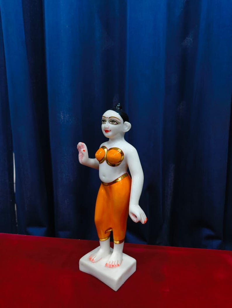 May include: A white and orange figurine of a person with black hair and red lips. The figurine is wearing an orange top and pants with gold trim. The figurine is standing on a white square base. The background is a blue curtain.