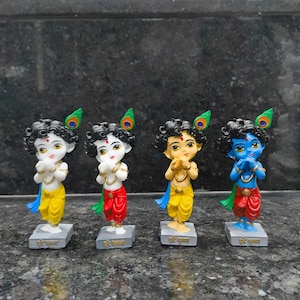 May include: Four small Krishna figurines. Each figurine has black curly hair, a peacock feather, and is wearing a dhoti. The figurines are in different colors: white, red, yellow, and blue. Each figurine is standing on a gray rectangular base.
