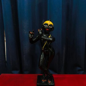 May include: A black statue of a deity with yellow and red accents. The figure is posed with hands raised in a gesture of blessing, standing on a black rectangular base. The background features a dark blue curtain and a red surface.