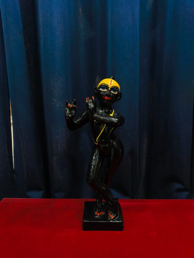 May include: A black statue of a deity with yellow and red accents. The figure is posed with hands raised in a gesture of blessing, standing on a black rectangular base. The background features a dark blue curtain and a red surface.