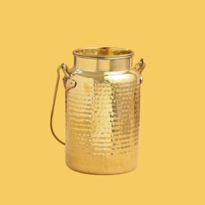 May include: A gold-colored metal milk can with a hammered texture. The can has a handle and a lid. The background is a solid yellow color. This decorative piece is suitable for home decor.