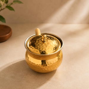 Op de afbeelding: Een kleine, goudkleurige container met een gehamerde textuur, met een deksel en een kleine houten handgreep. De container heeft een zilveren rand en een afgeronde vorm. De achtergrond is neutraal beige.