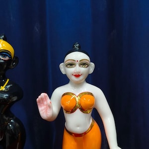 May include: A white and orange figurine of a woman with a serene expression. The figurine has orange clothing with gold accents and a small red dot on the forehead. A black figurine is partially visible on the left.