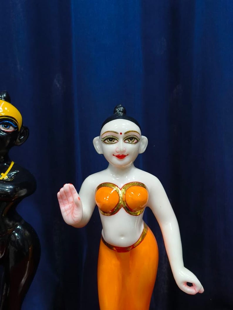 May include: A white and orange figurine of a woman with a serene expression. The figurine has orange clothing with gold accents and a small red dot on the forehead. A black figurine is partially visible on the left.