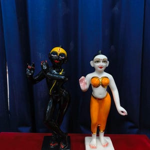 May include: Two statues of deities. One is black with yellow accents, the other is white with orange and gold accents. Both are standing on square bases. The background is a dark blue curtain and a red surface.
