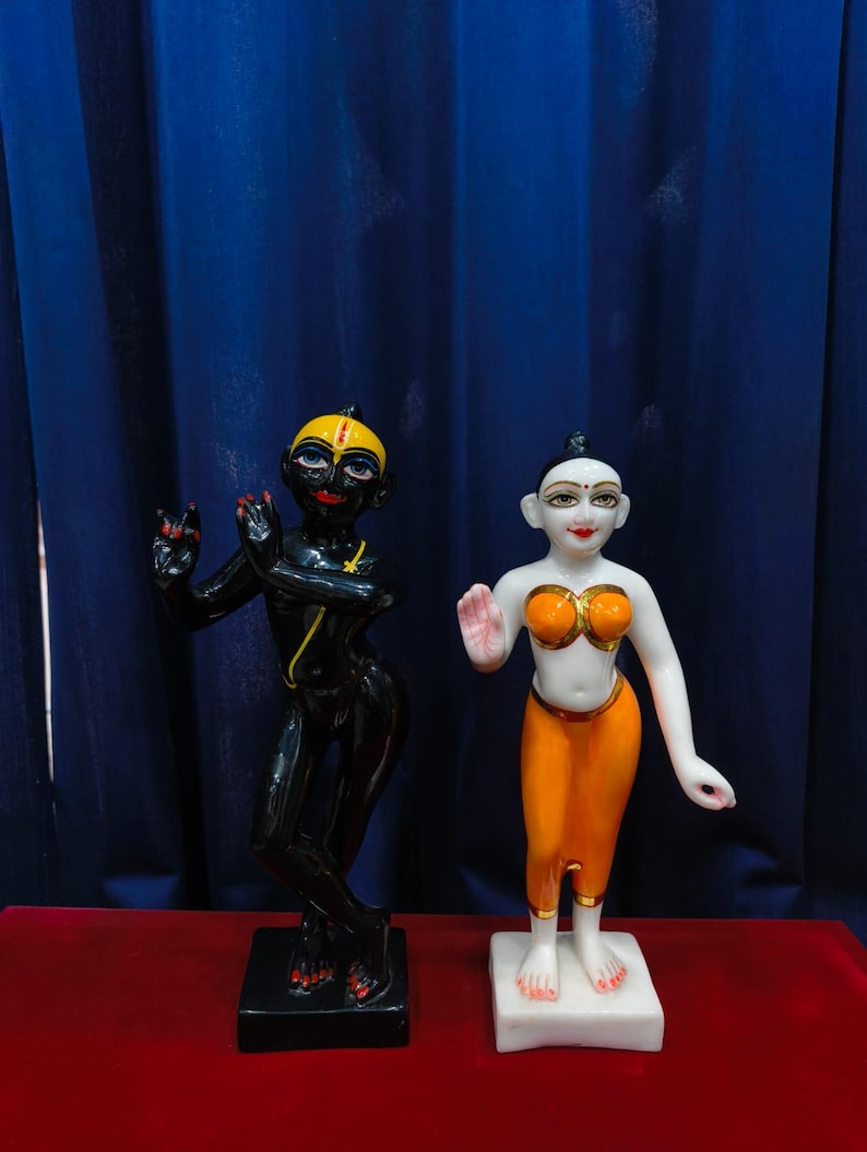 May include: Two statues of deities. One is black with yellow accents, the other is white with orange and gold accents. Both are standing on square bases. The background is a dark blue curtain and a red surface.