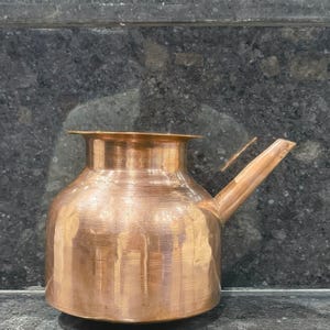 May include: A copper-colored metal pitcher with a rounded body, a wide mouth, and a spout extending from the side. The pitcher has a textured surface and sits on a dark, speckled surface. The design is simple and functional.