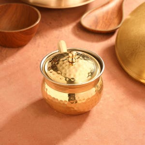 May include: A small, hammered gold-colored container with a lid and a small wooden spoon. The container is round with a textured surface. Other wooden and gold serving pieces are in the background.
