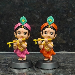 Könnte beinhalten: Zwei dekorative Figuren eines jungen Mannes, der Flöte spielt. Eine Figur trägt ein rosa Outfit und einen Turban, die andere ein orangefarbenes Outfit und einen Turban. Beide haben goldenen Schmuck und eine grüne Feder. Sie stehen auf schwarzen Sockeln.