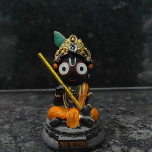 Könnte beinhalten: Kleine, schwarze Figur einer Gottheit mit großen weißen Augen, einem goldenen Kronen und einem gelben Stab. Die Gottheit sitzt auf einem grauen Sockel mit goldenen Akzenten und orangefarbener Kleidung. Der Sockel trägt Text in einer Fremdsprache.