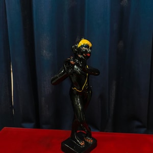 May include: A black figurine with a yellow head covering and red lips. The figurine is standing on a black base and has a gold-colored necklace. The background is a dark blue curtain and a red surface.