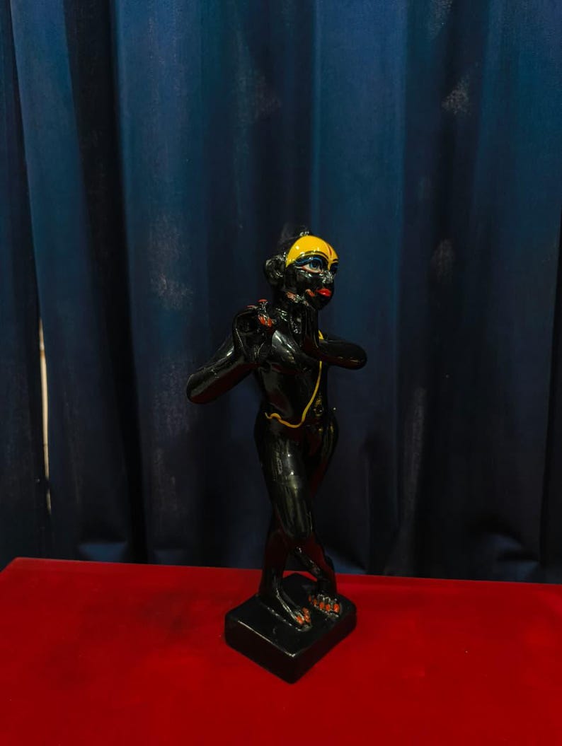 May include: A black figurine with a yellow head covering and red lips. The figurine is standing on a black base and has a gold-colored necklace. The background is a dark blue curtain and a red surface.