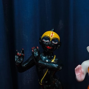 May include: A black figurine of a deity with a yellow head, blue eyes, and red lips and fingernails. The figurine has a yellow necklace and is posed with hands raised. The background is a dark blue curtain.
