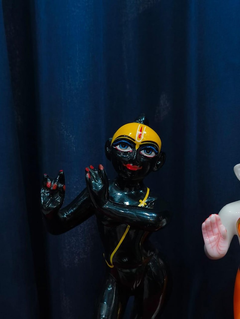 May include: A black figurine of a deity with a yellow head, blue eyes, and red lips and fingernails. The figurine has a yellow necklace and is posed with hands raised. The background is a dark blue curtain.
