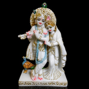 May include: A white marble statue depicting two figures with gold detailing. One figure holds a flute, the other is draped in white. A peacock is at the base. The statue is set against a black background. The statue is likely a religious or cultural item.