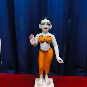 May include: A white statue of a figure with orange clothing and gold accents. The figure is standing on a white base, with one hand raised. The background is a blue curtain and a red surface.