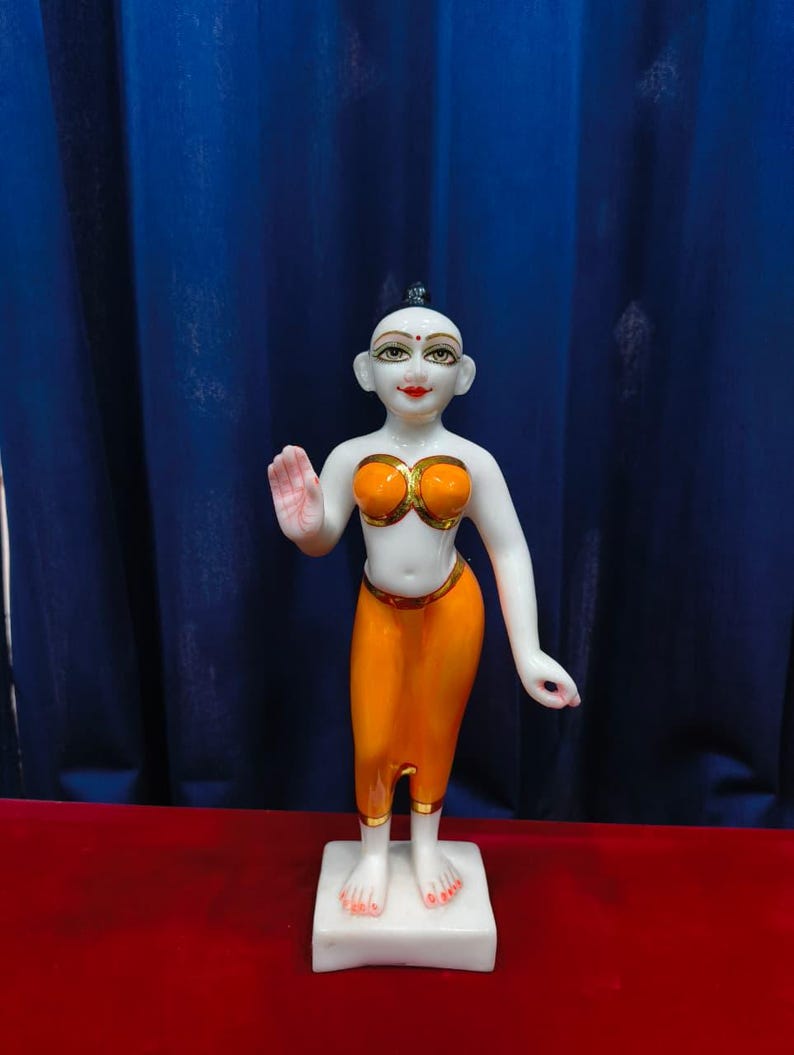 May include: A white statue of a figure with orange clothing and gold accents. The figure is standing on a white base, with one hand raised. The background is a blue curtain and a red surface.