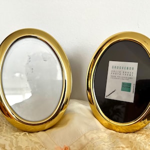 May include: Two oval gold photo frames. One has a white interior, the other a black interior with a label reading "GROSVENOR SOLID BRASS PHOTO FRAME". The frames are approximately 12cm x 18cm.