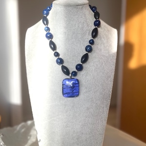 May include: A necklace featuring a rectangular blue glass pendant with horizontal black lines, suspended from a strand of alternating round blue marbled beads, elongated black beads, and small silver-toned spacers on a grey display bust.